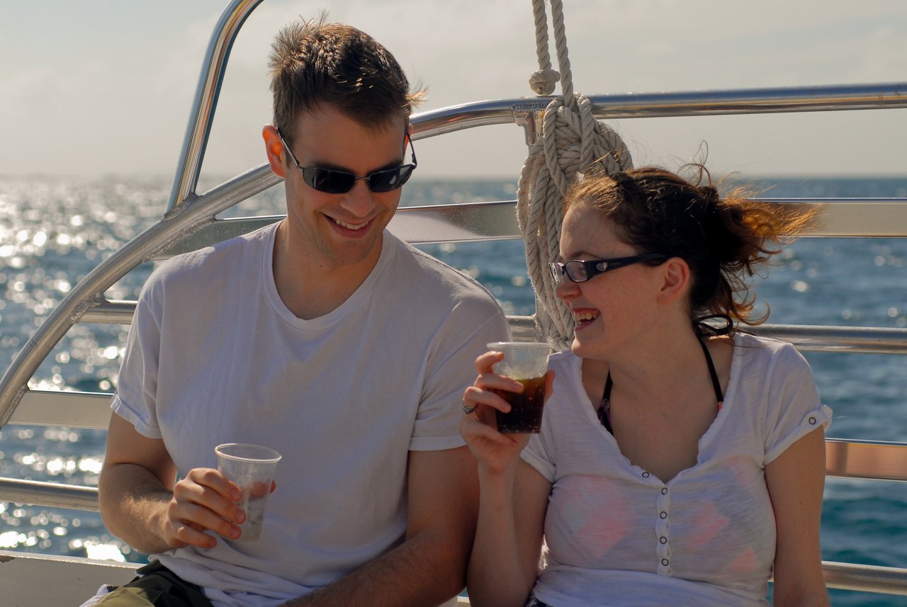 A man and woman sit on a boat, smiling and holding drinks while looking at each other.