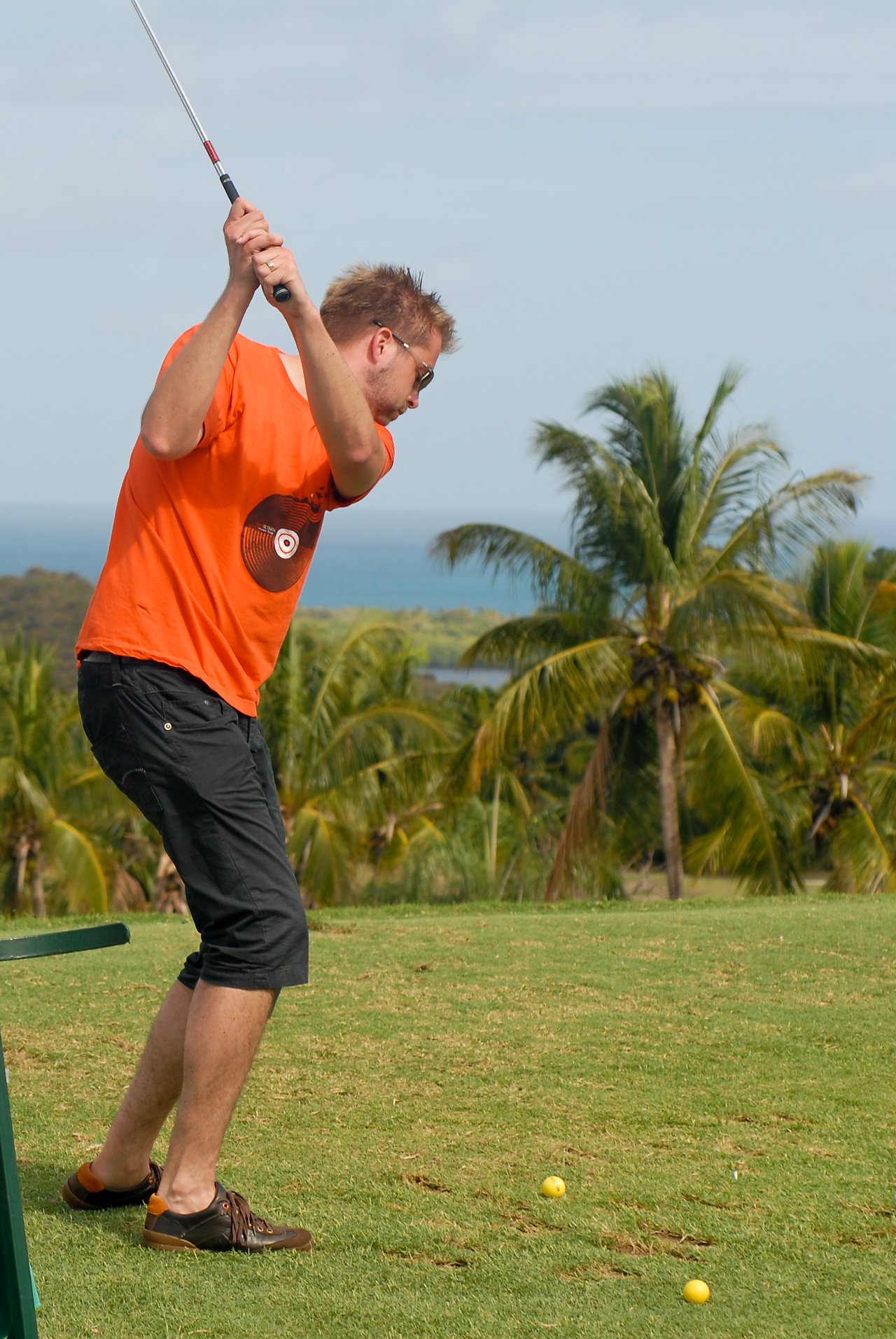 A man in an orange shirt practices his golf swing on a grassy course with golf balls nearby.