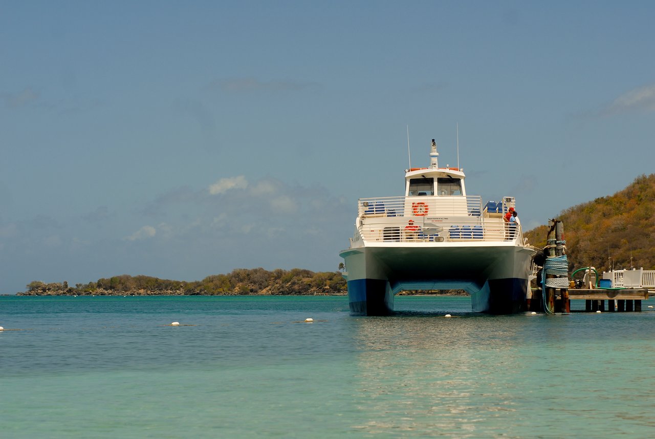 A white ferry is docked at a pier in clear turquoise water, with a small island in the background.