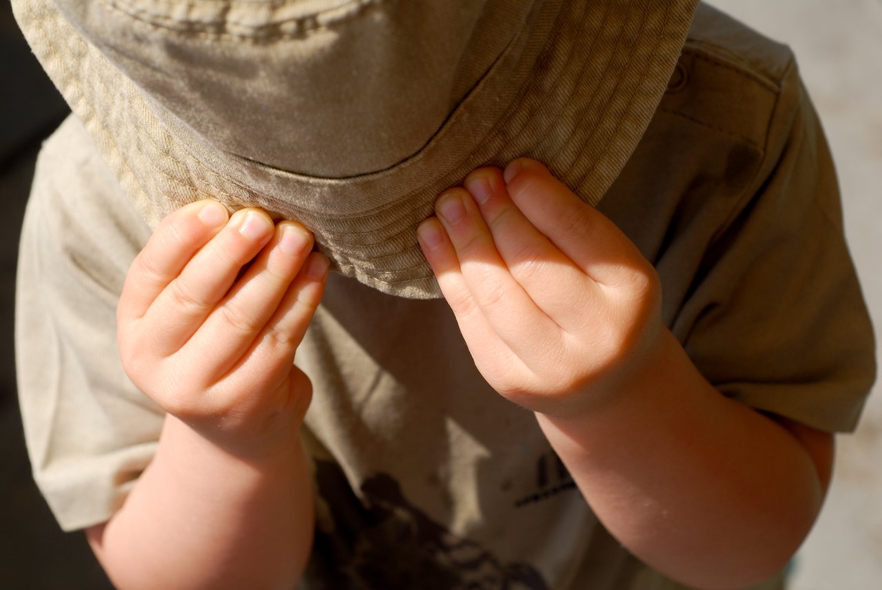 A child wearing a hat holds the brim with both hands, partially covering their face.