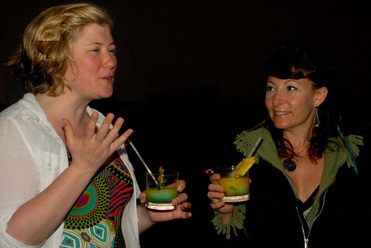 Two women at a party holding drinks, one speaking with hand gestures while the other listens and smiles.