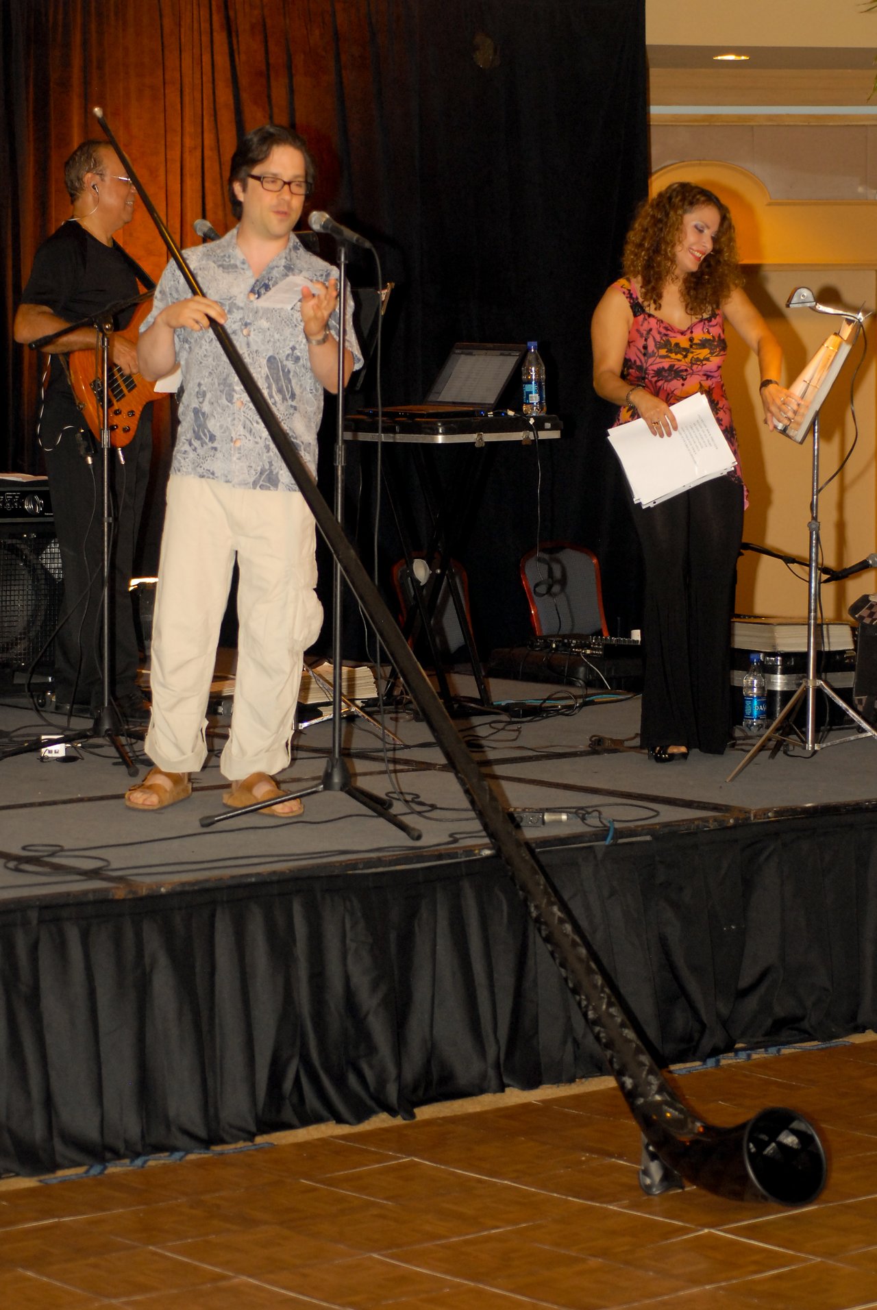 A man on stage holds an alphorn while speaking into a microphone, with musicians performing in the background.