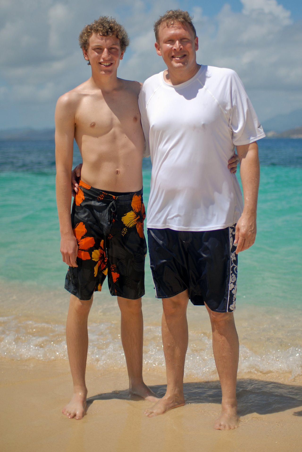 Two people stand side by side on a sandy beach, smiling at the camera with ocean waves behind them.