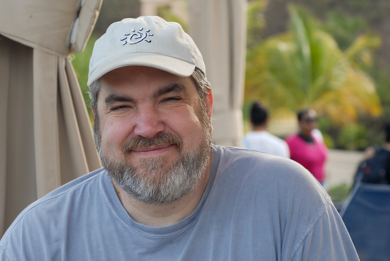 A bearded man in a gray shirt and white cap smiles at the camera, with people and palm trees behind him.