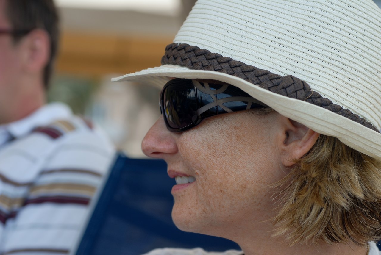 A woman wearing a sun hat and sunglasses smiles while sitting outdoors.