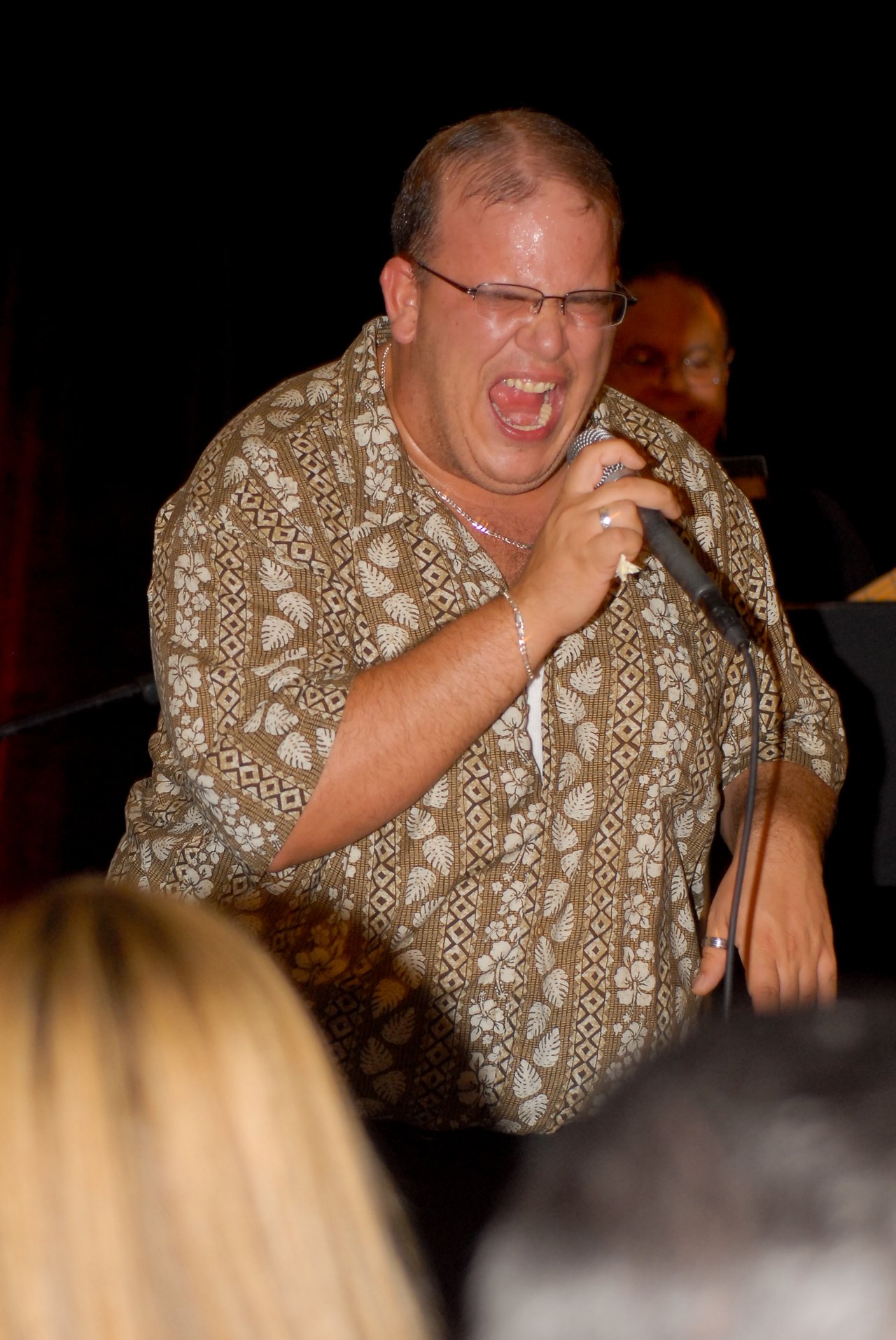 A man in a patterned shirt sings passionately into a microphone during a live performance, with an audience watching.