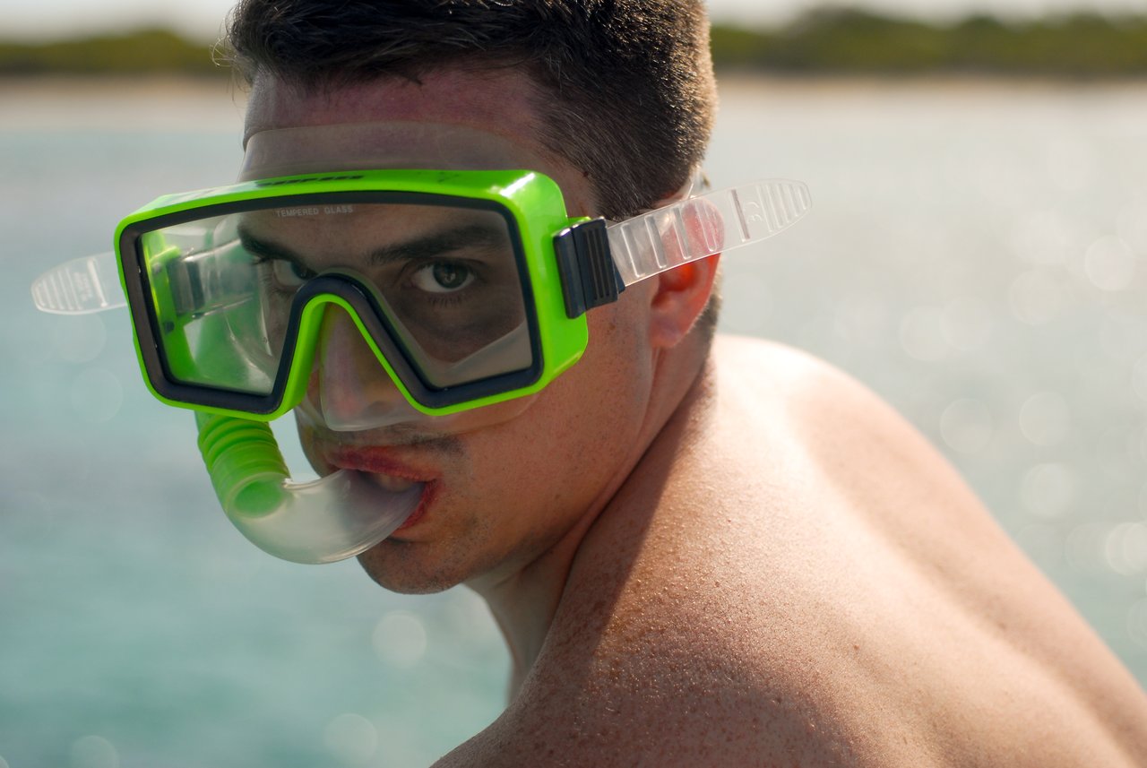 A person wearing a green snorkel mask looks toward the camera with water and shoreline in the background.