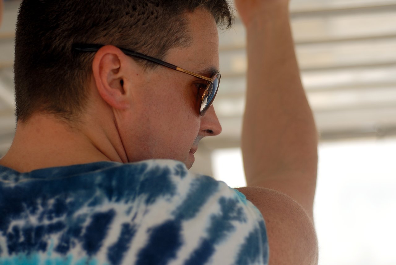 A man wearing sunglasses and a tie-dye shirt holds onto a railing while looking to the side.