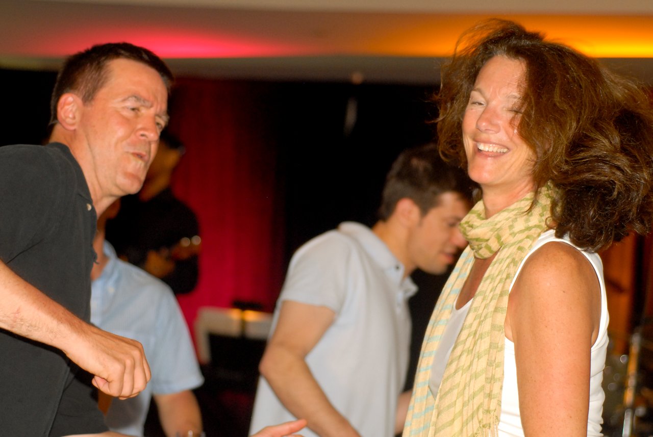 A woman with curly hair and a scarf smiles while dancing, surrounded by other people enjoying the music.