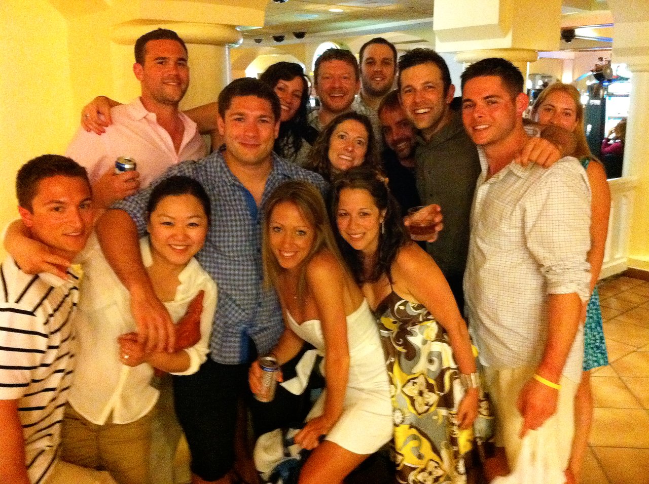 A group of friends posing together at a party, smiling and holding drinks in a lively indoor setting.
