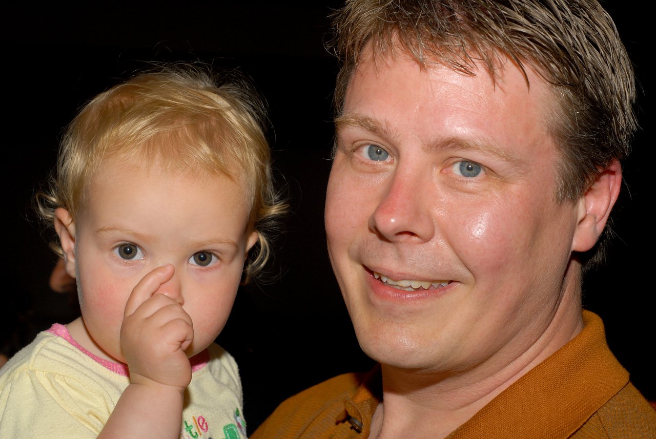 A man smiles at the camera while holding a young child who is sucking their thumb.