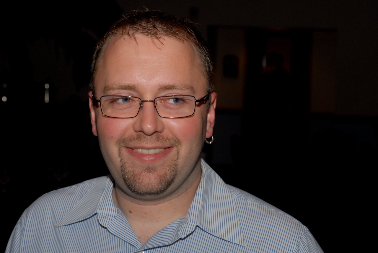 A man wearing glasses and a striped shirt smiles while looking slightly to the side in a dimly lit setting.