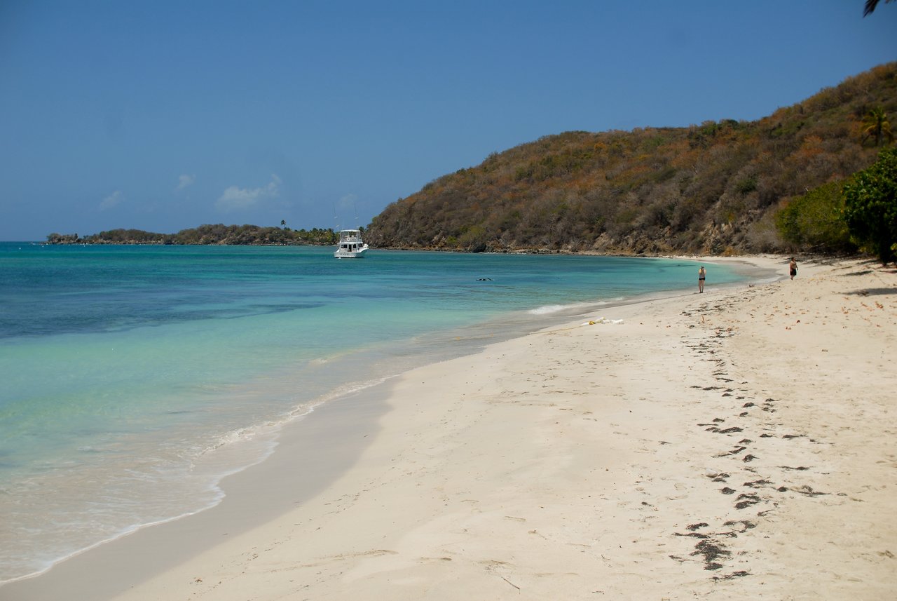 A sandy beach with clear blue water, a boat anchored nearby, and two people walking along the shore.