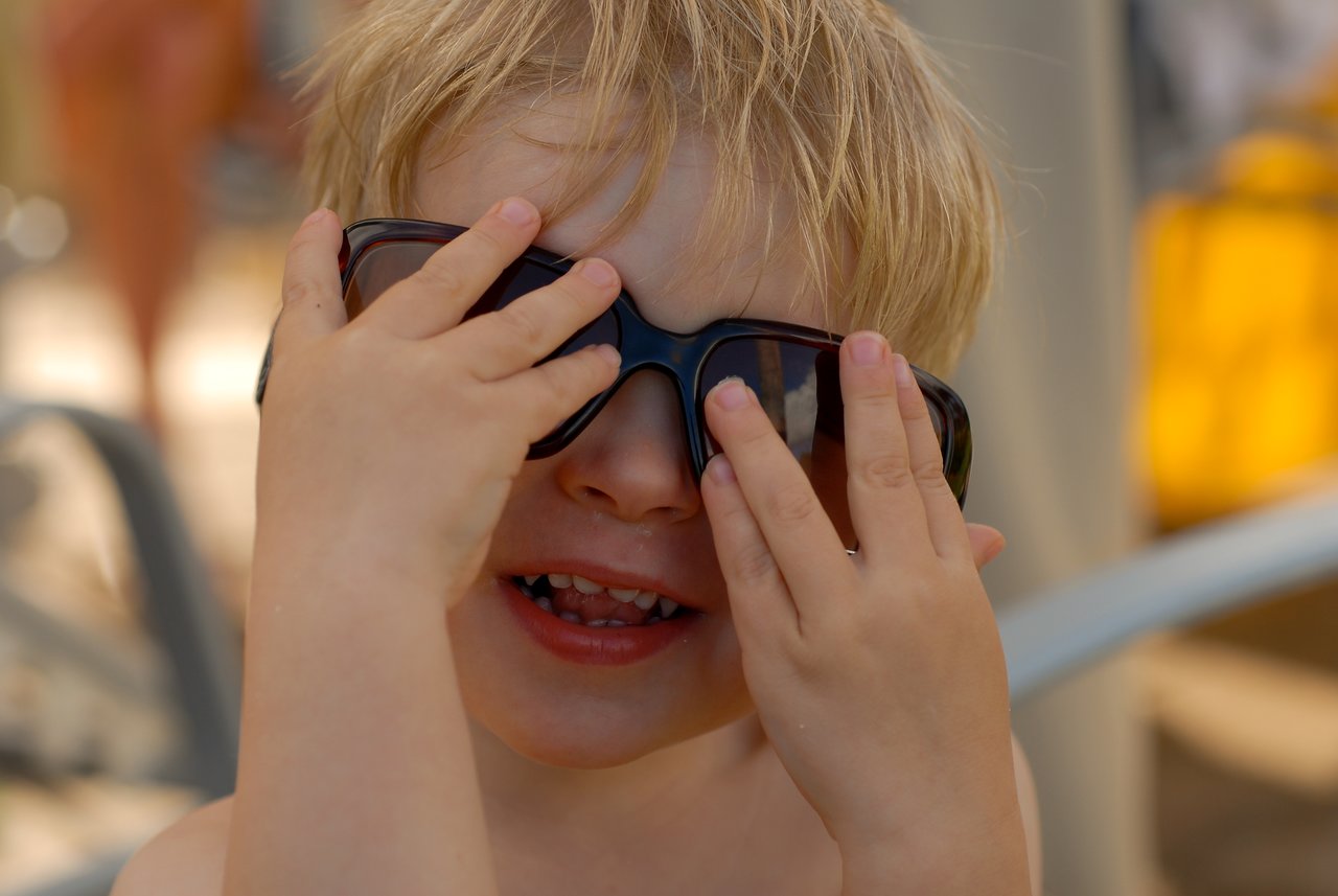 A young child with blonde hair playfully adjusts oversized sunglasses while smiling.
