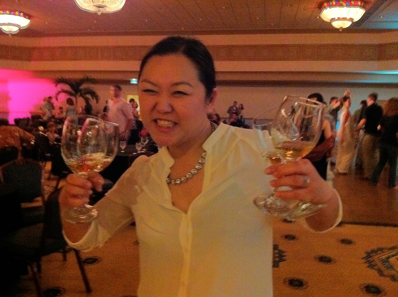 A woman in a white blouse smiles while holding two glasses with drinks at a lively indoor event.