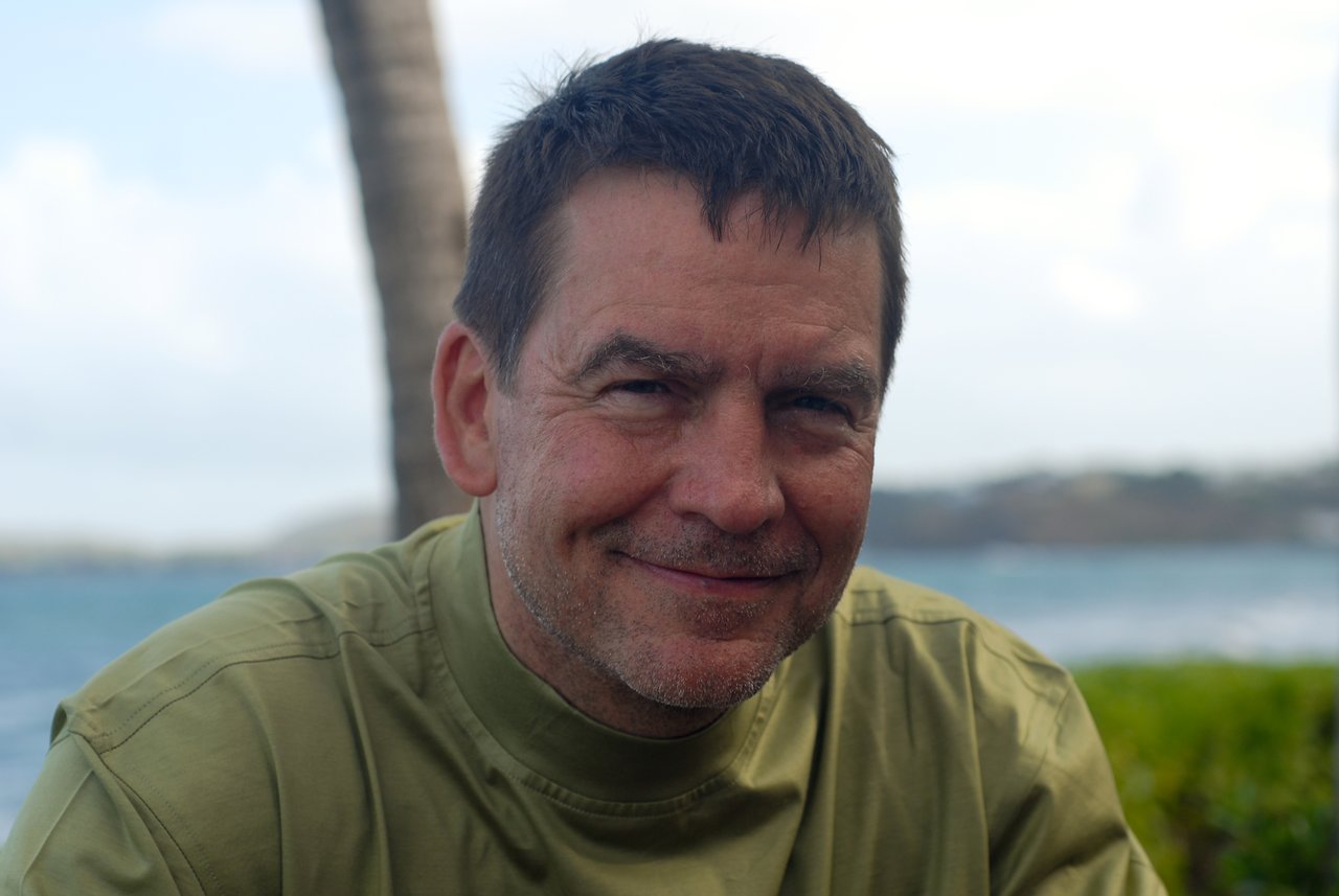 A man in a green shirt smiles at the camera with a blurred ocean and palm tree in the background.