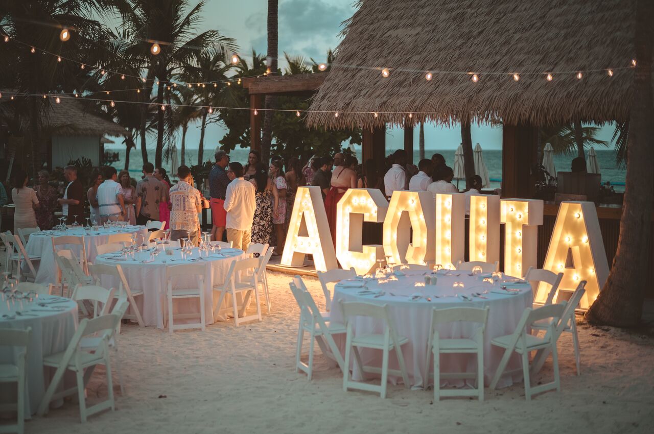 Large illuminated letters spelling "Acquia" are displayed next to tables arranged for a beachside dinner.