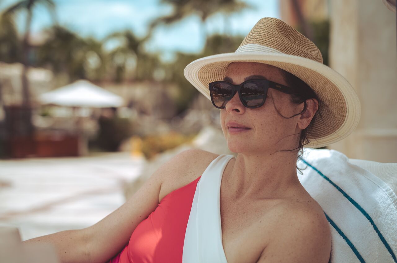 Woman in a sun hat and sunglasses relaxes on a lounge chair by the pool.