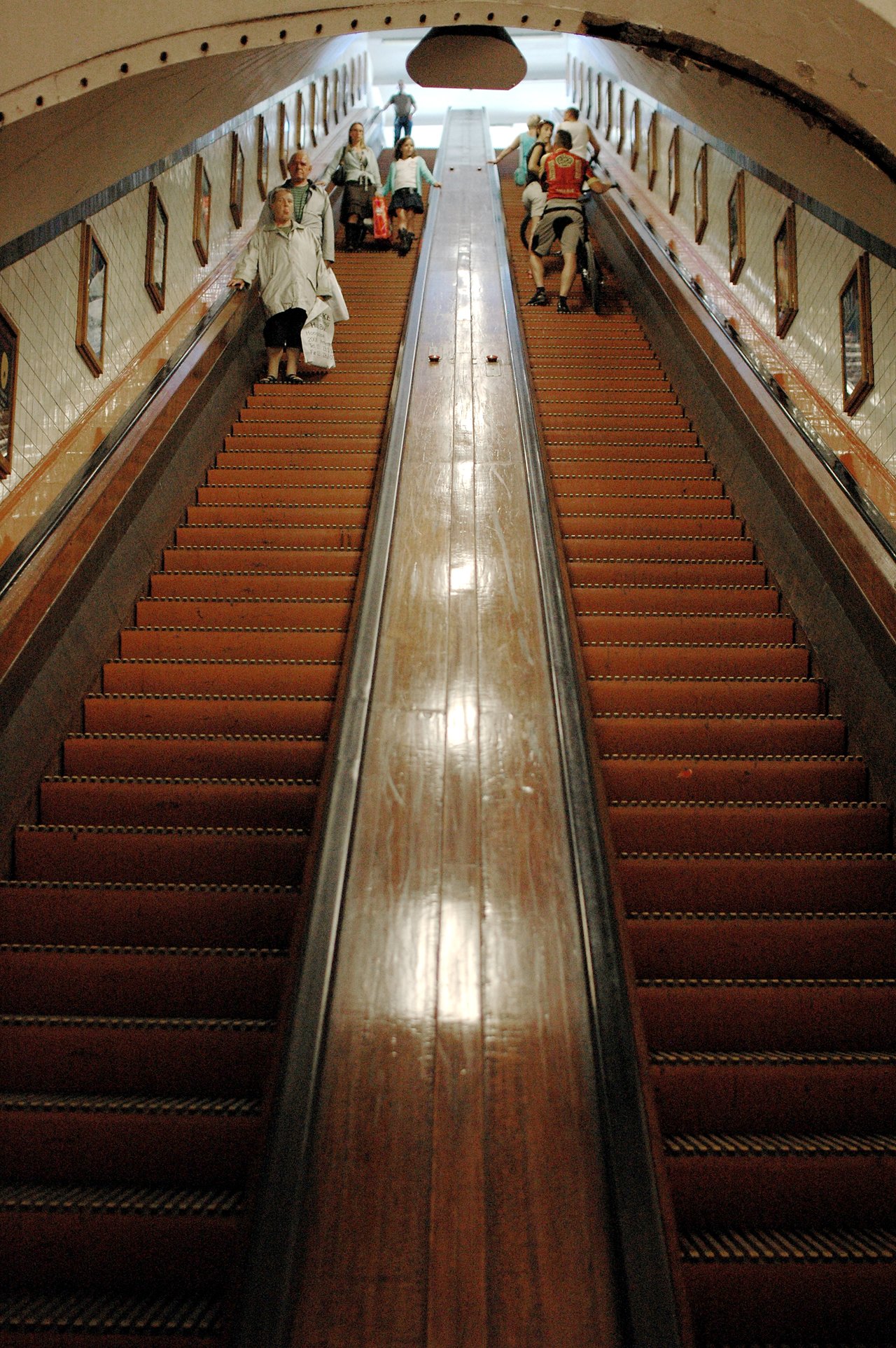 People walk up and down a long wooden escalator in an underground passage, carrying bags and backpacks.