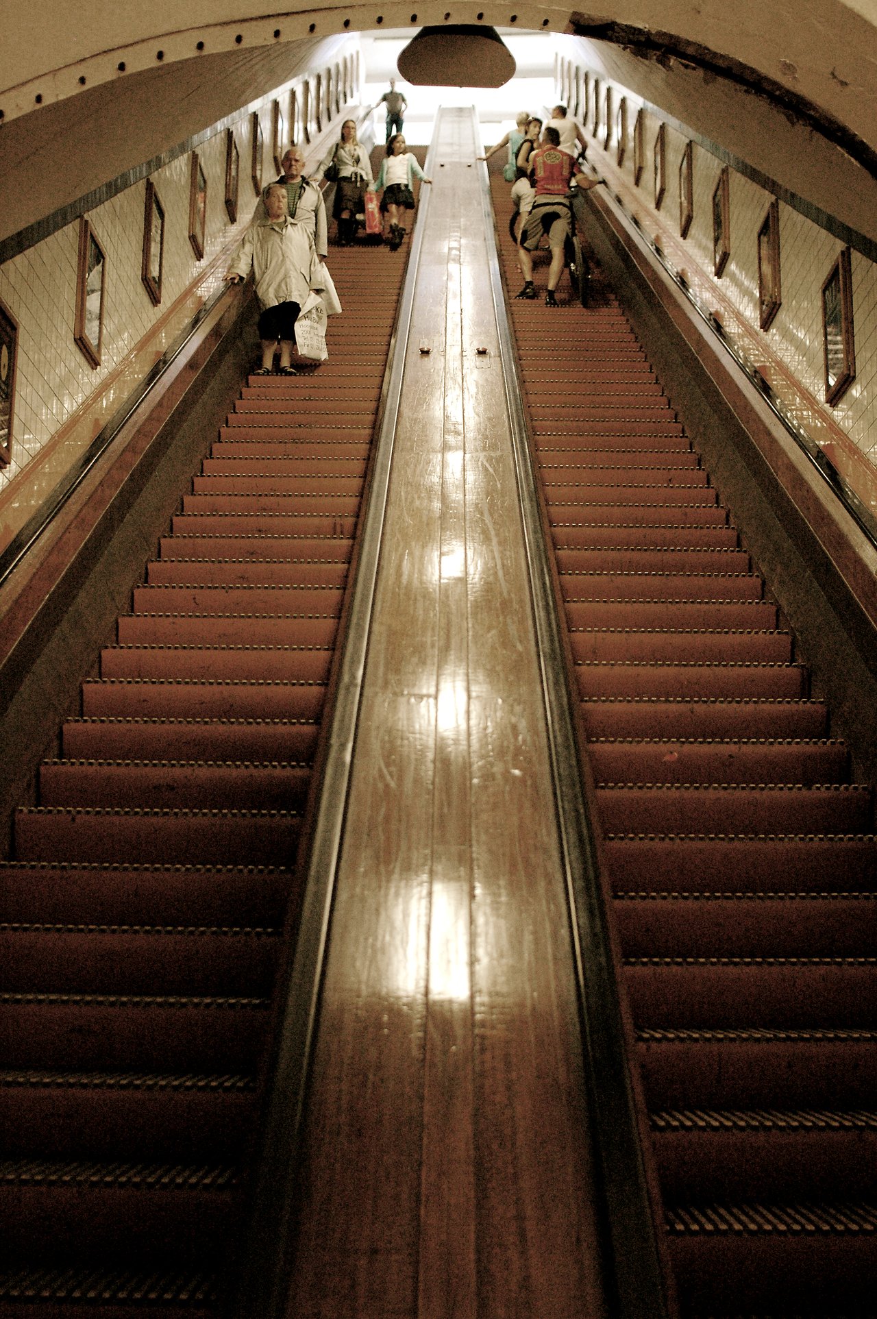 People walk up and down a long wooden escalator in an underground passage with framed pictures on the walls.