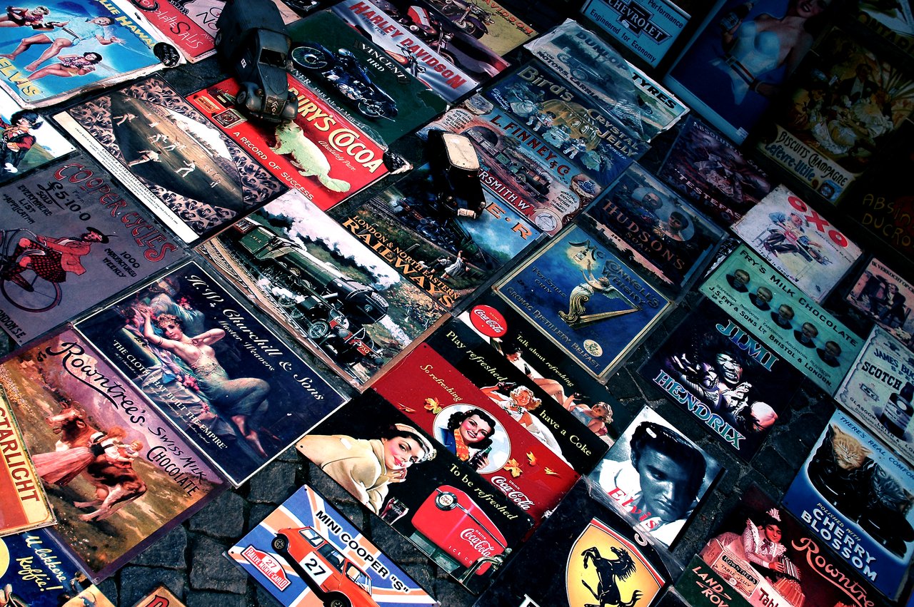 A collection of vintage metal signs with various advertisements and images displayed on a cobblestone surface.