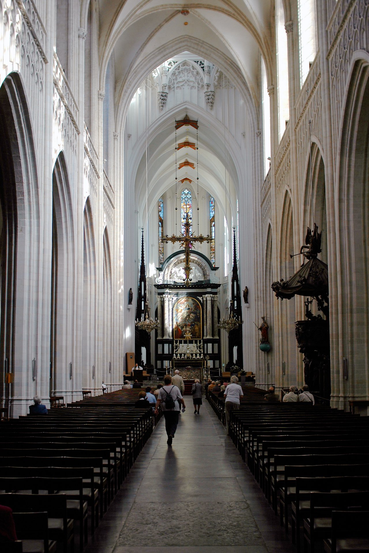 People walk down the central aisle of a large church toward the ornate altar, surrounded by tall arches and pews.