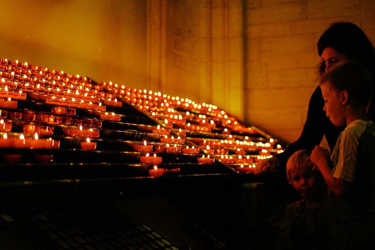 A woman and two children stand near rows of lit candles, appearing to light or observe them.