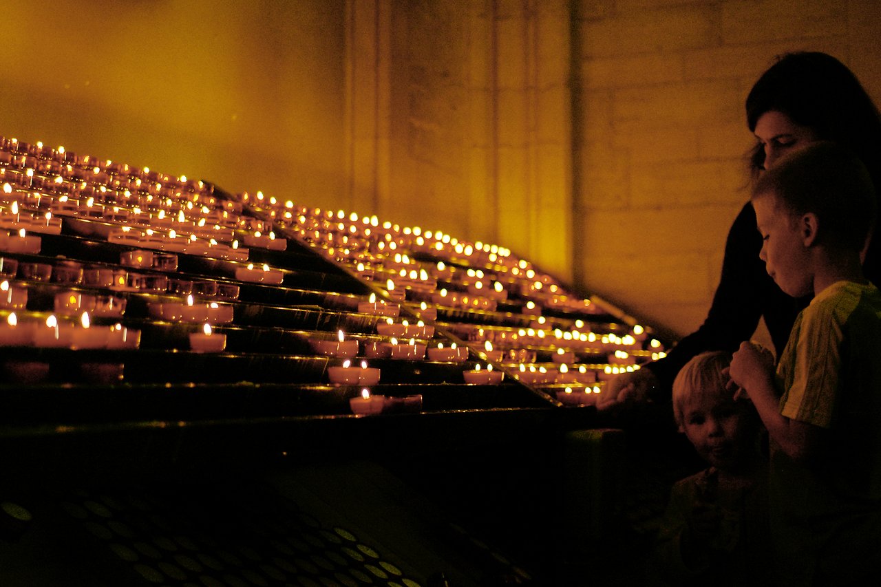 A woman and two children stand near rows of lit candles, appearing to observe or participate in a ritual.
