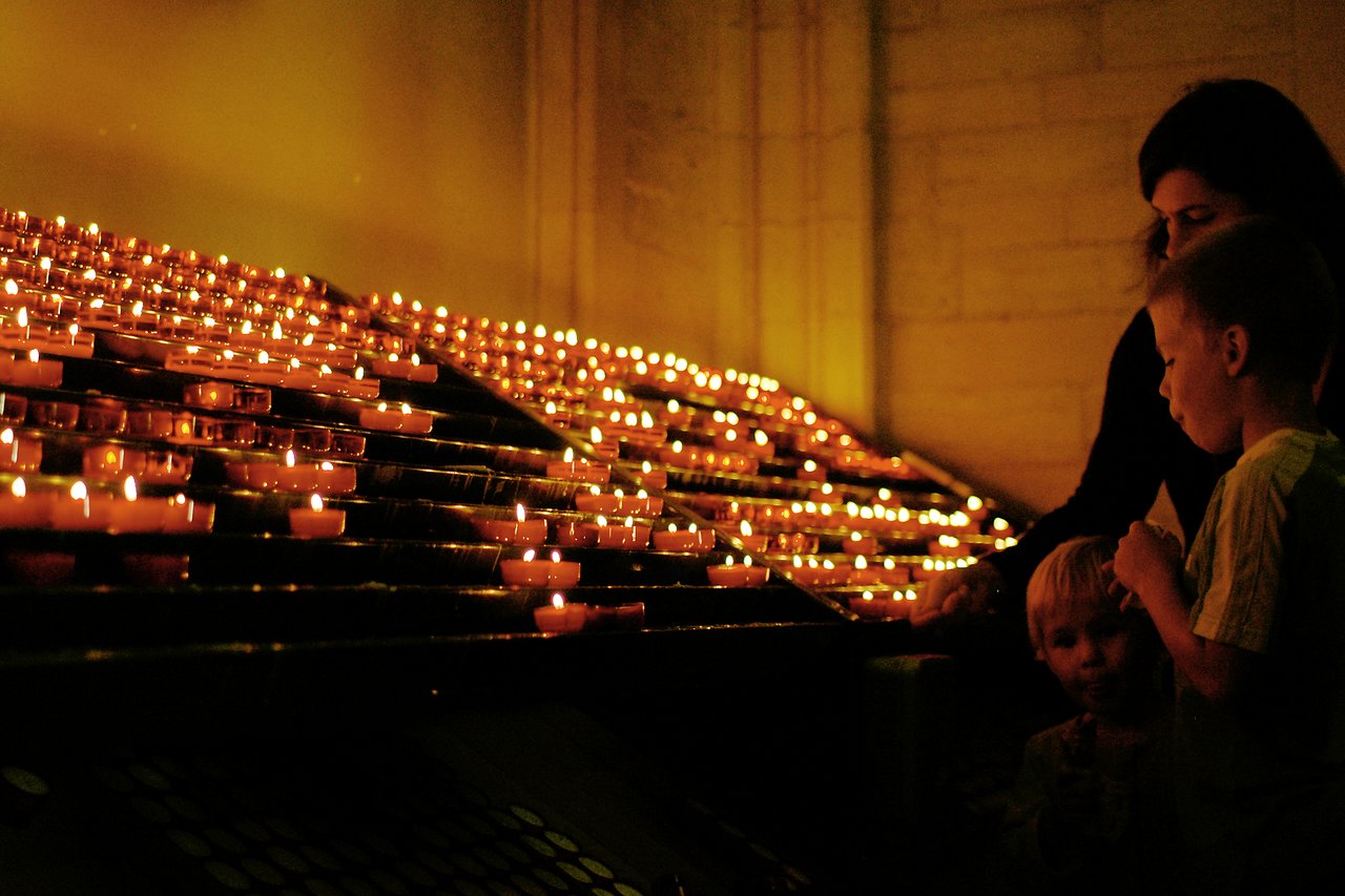A woman and two children stand near a display of lit candles in a dimly lit space.
