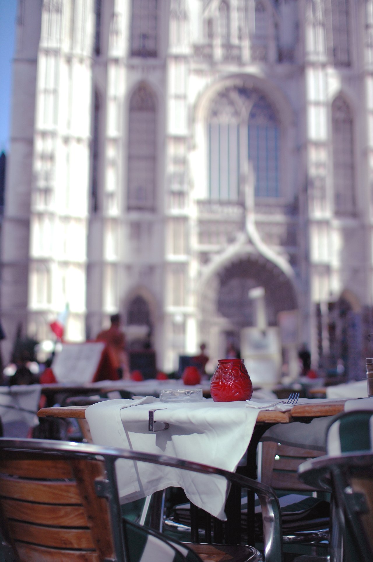 Outdoor café table with a white tablecloth, red candle holder, and utensils, set against a blurred historic building.