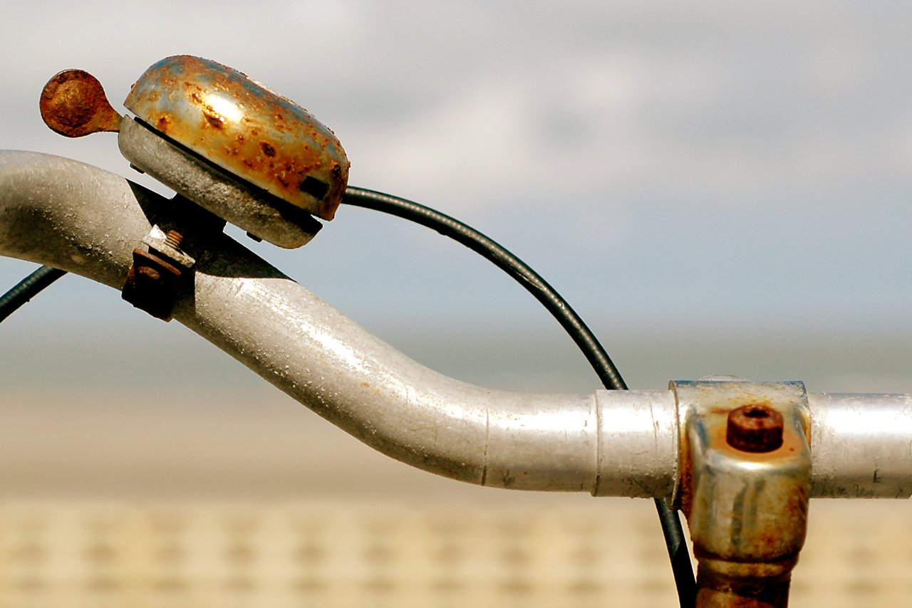 Close-up of a rusty bicycle handlebar with a worn bell and cables against a blurred outdoor background.