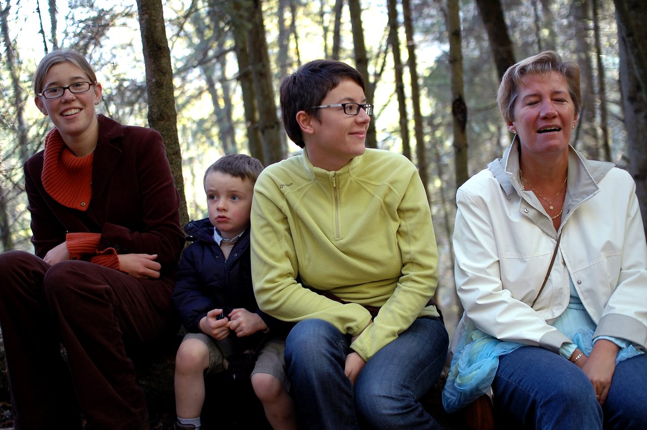 Four people sit on a bench in a wooded area, engaged in conversation.