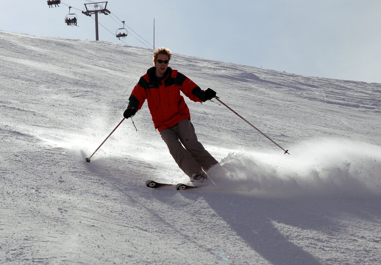 A person in a red jacket skis downhill, kicking up snow while using ski poles for balance.