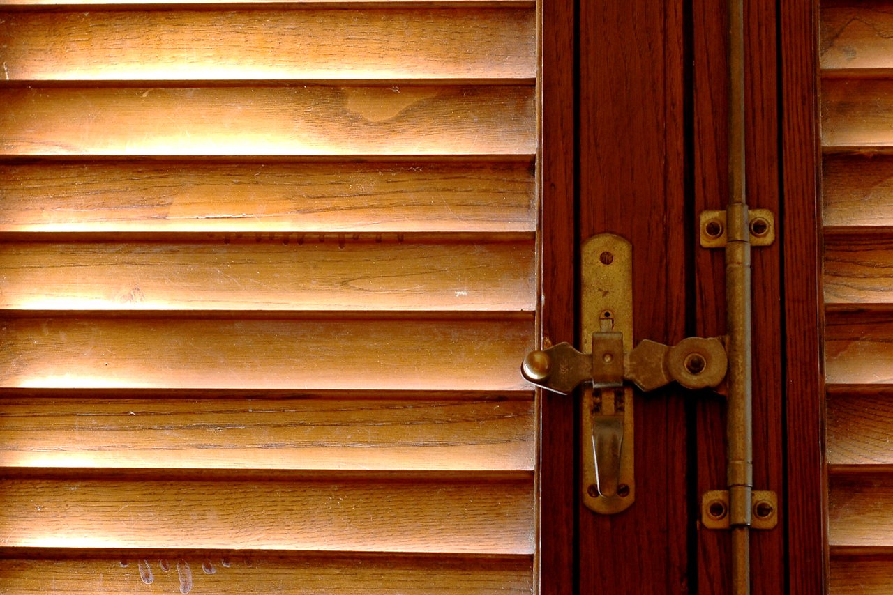 Sunlight filters through wooden shutters, casting a warm glow on a closed window with a brass latch.