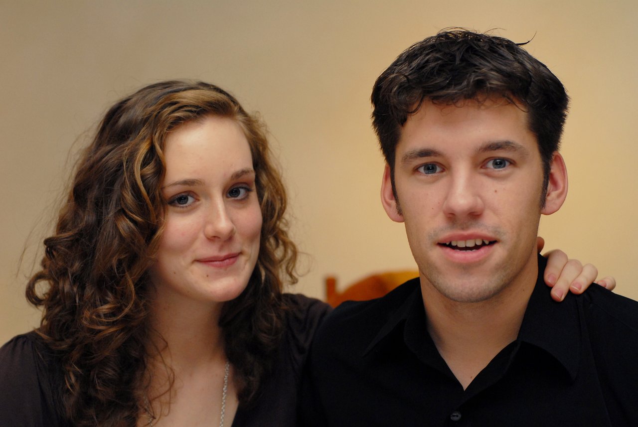 A woman and a man in black shirts pose together, with the woman resting her hand on his shoulder.