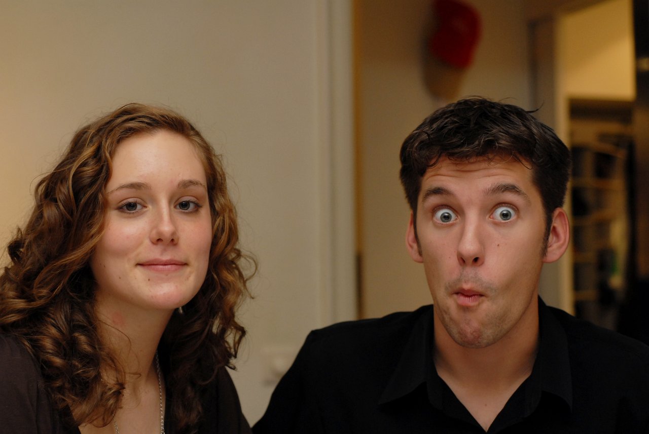 A woman smiles slightly while a man next to her makes a surprised expression with wide eyes and pursed lips.