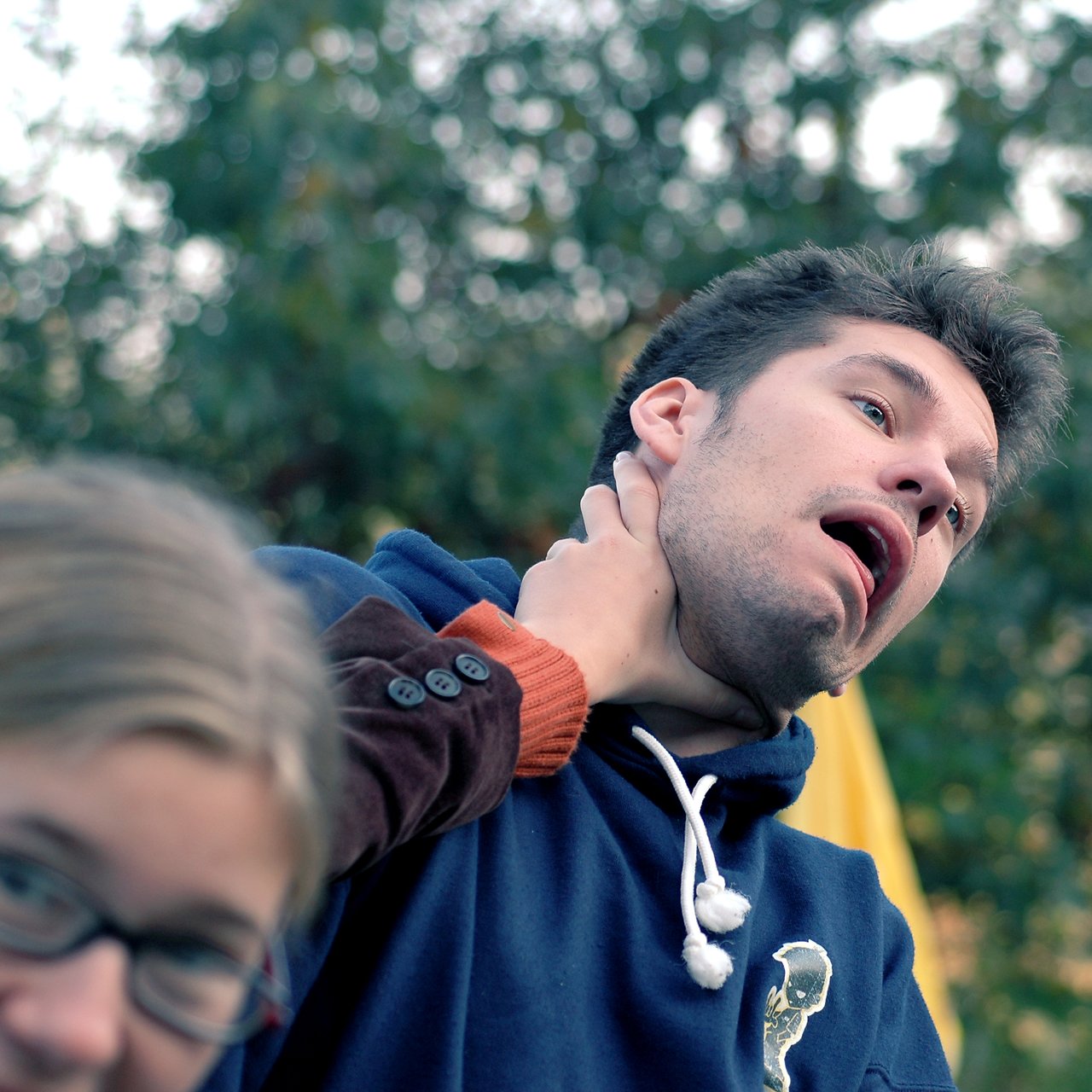A person in a blue hoodie reacts dramatically while another person's hand grabs their neck playfully.