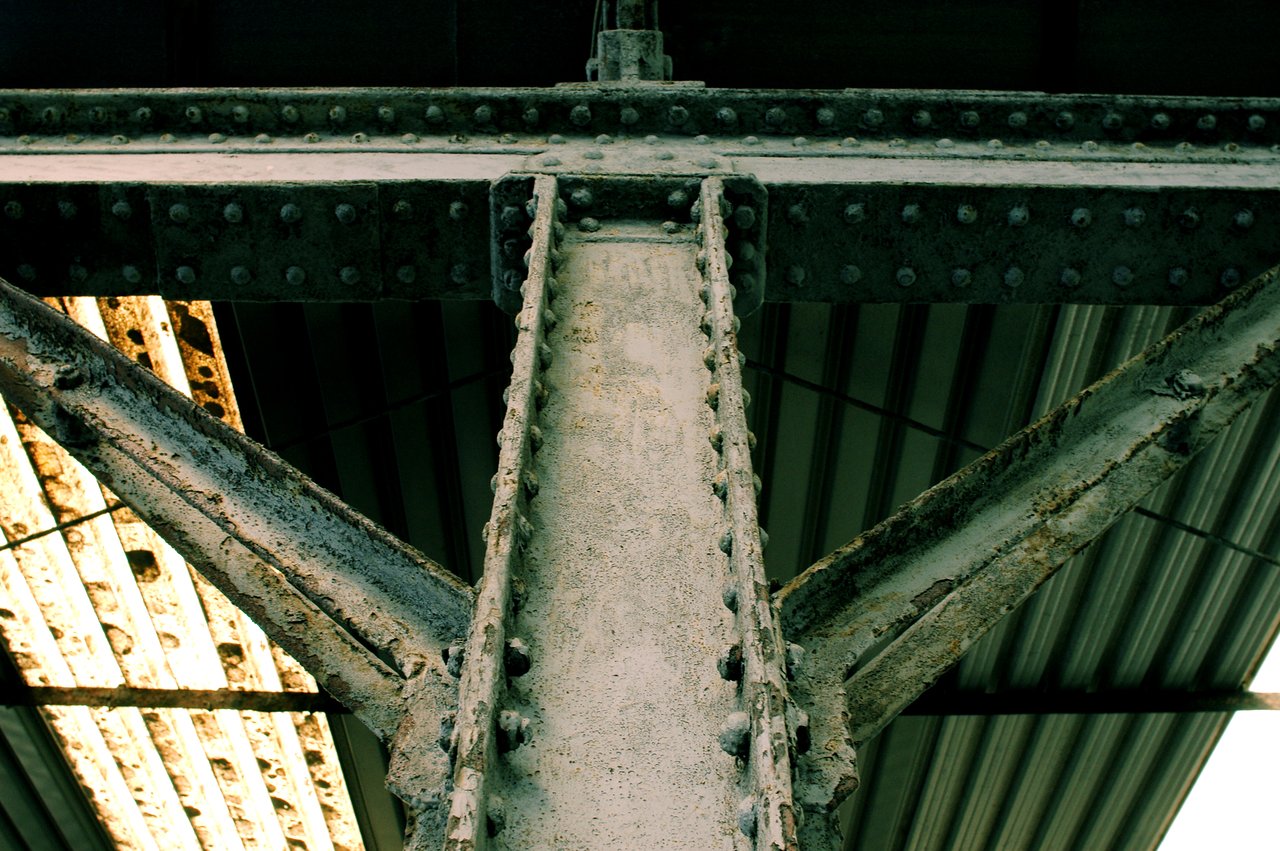 Close-up of a rusted metal beam with rivets, supporting a corrugated metal roof in an industrial structure.