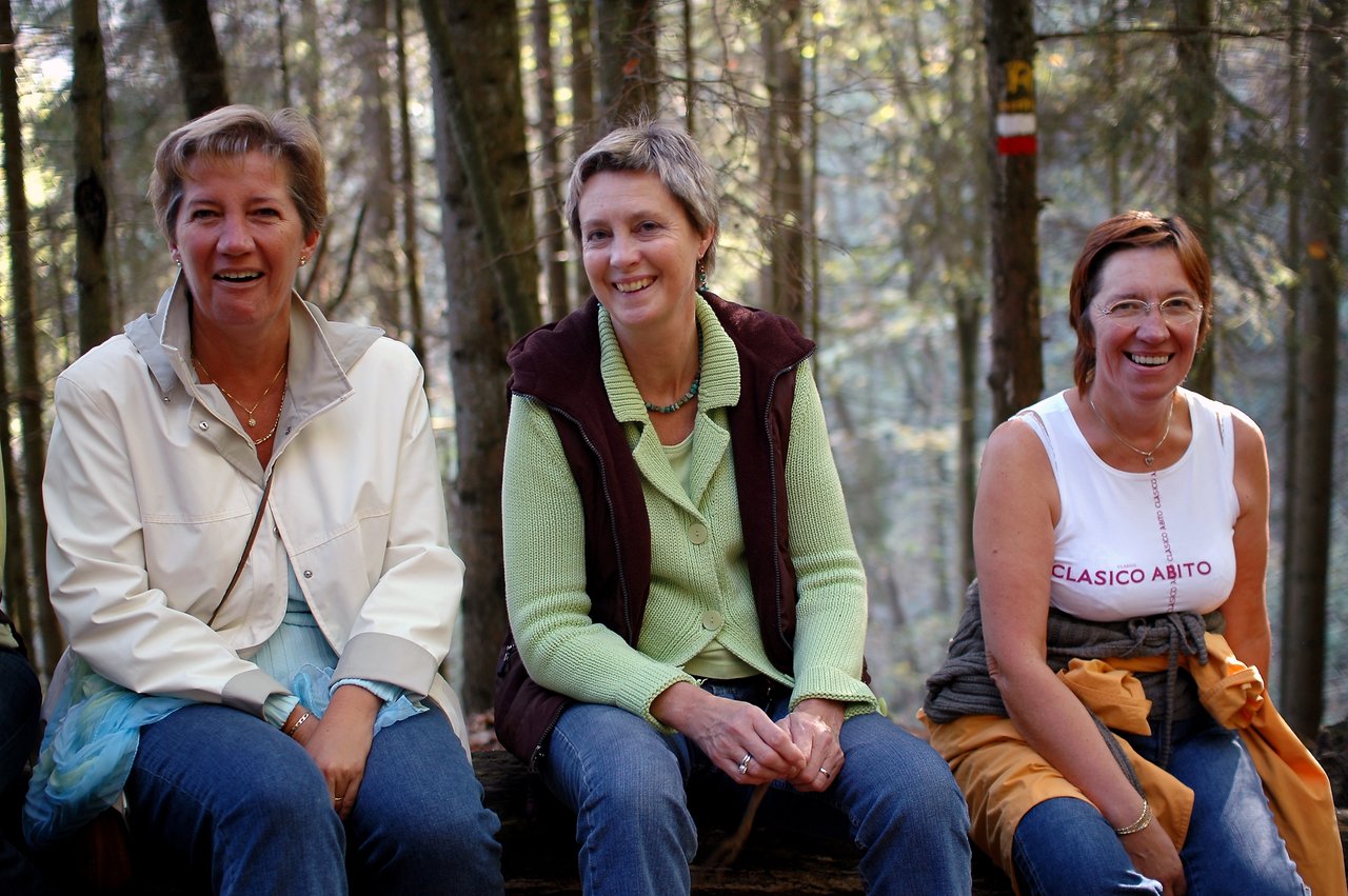 Three women sitting on a bench in a forest, smiling and looking at the camera.