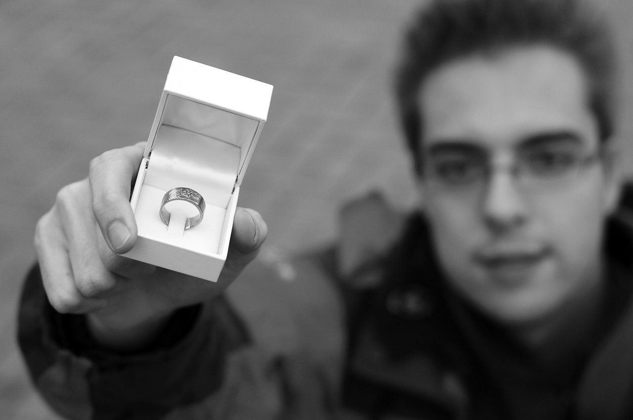 A person holds out an open ring box with a ring inside, suggesting a marriage proposal.