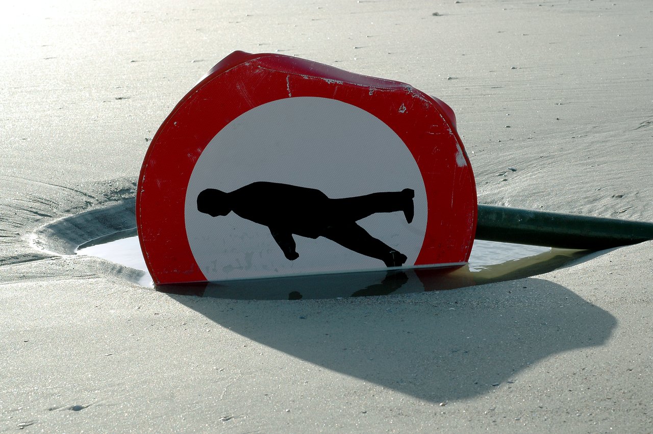 A partially submerged traffic sign with a tilted pedestrian symbol, appearing distorted due to its position in the sand.