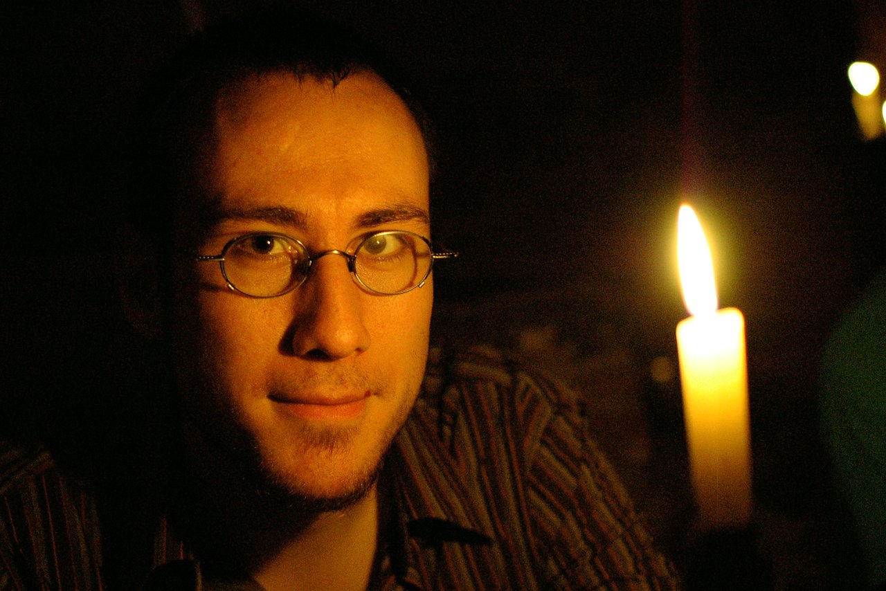 A person wearing glasses sits in dim lighting with a lit candle nearby.