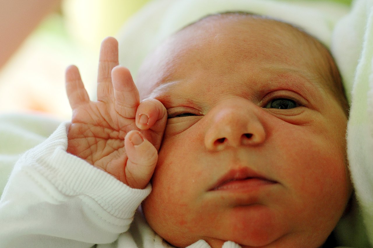 A newborn baby lies on a soft surface, touching their face with a tiny hand while gazing slightly upward.