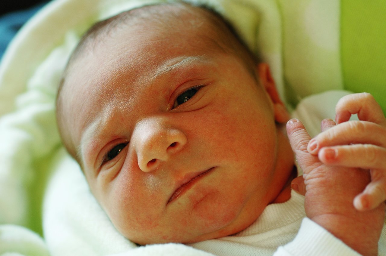 A newborn baby lies on a soft blanket, gazing slightly upward with a calm expression and curled fingers.