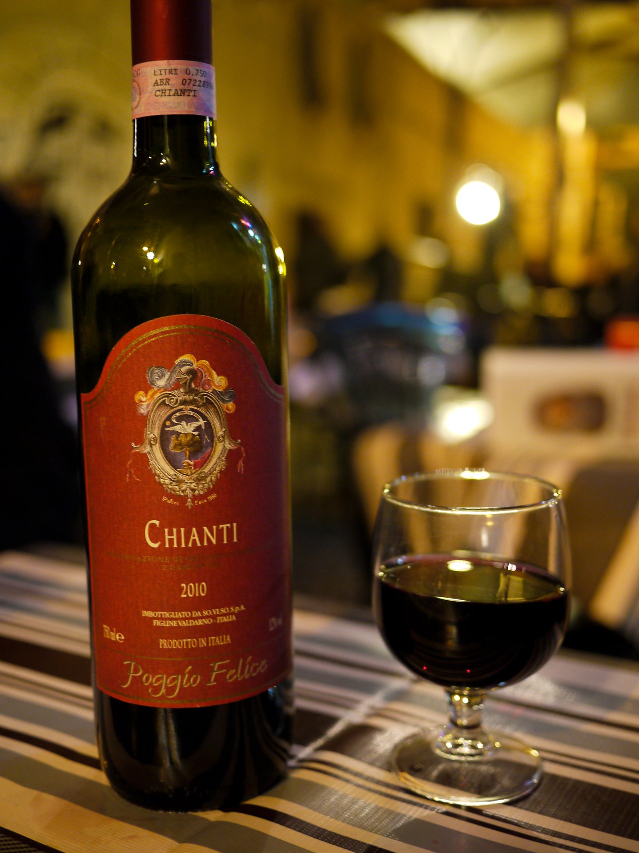 A bottle of Chianti wine and a glass filled with red wine on a striped tablecloth.