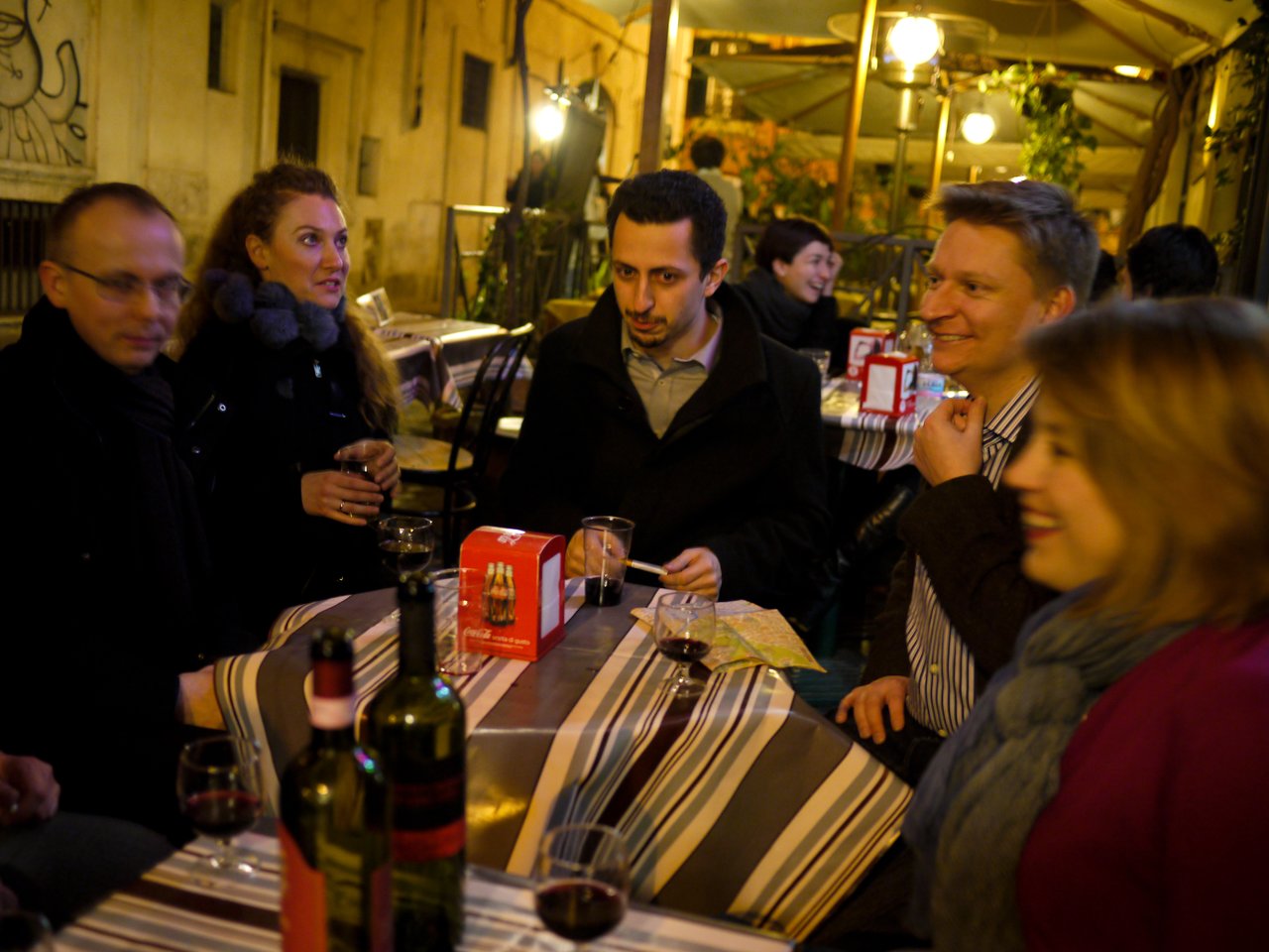 A group of people sits at an outdoor restaurant table, drinking wine and talking in a warmly lit setting.