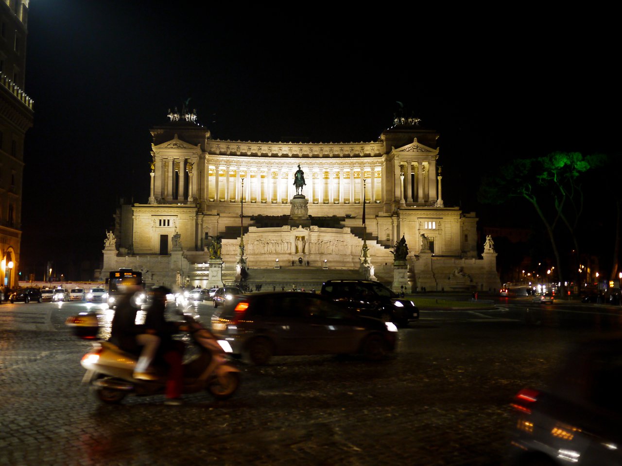 Busy nighttime street in Rome with cars and a scooter passing in front of an illuminated historic building.