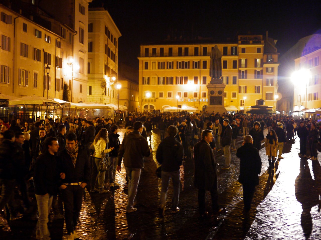 A crowded city square at night with people walking and talking near a statue and illuminated buildings.