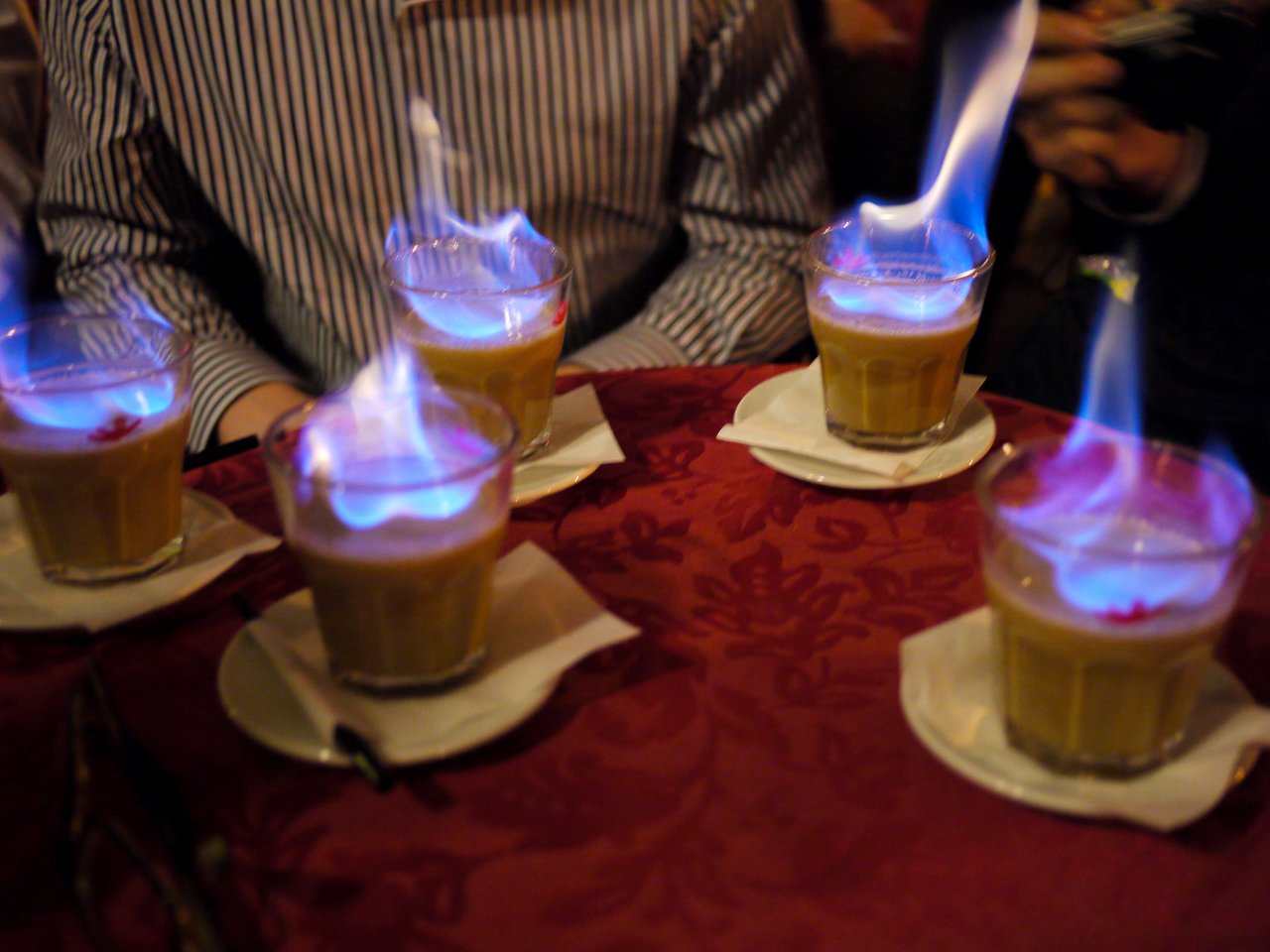 Five glasses of flaming drinks on a red tablecloth, with people sitting nearby.