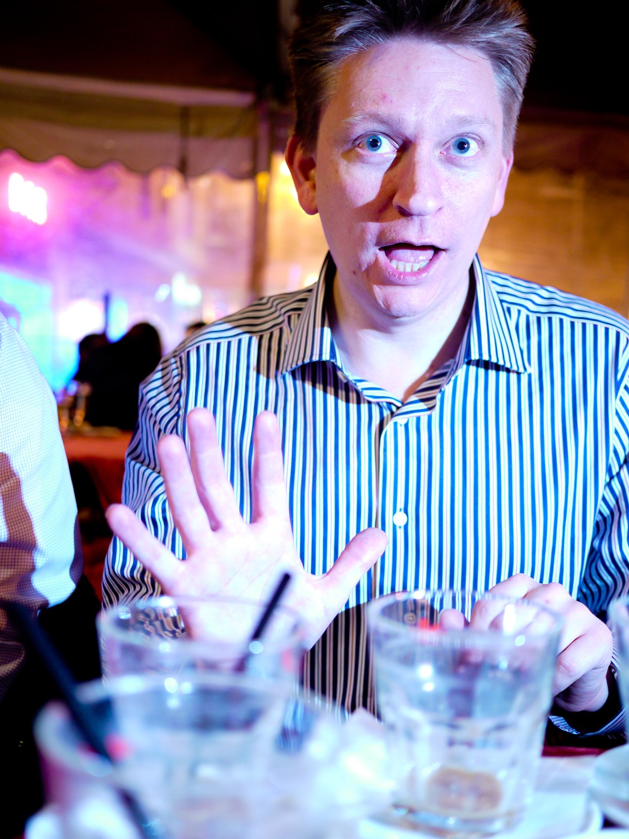 A man in a striped shirt raises his hand and reacts with surprise at a table with drinks.